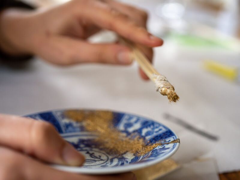 A close up image of a hand holding a soft brush with gold dust on it above a blue pottery plate